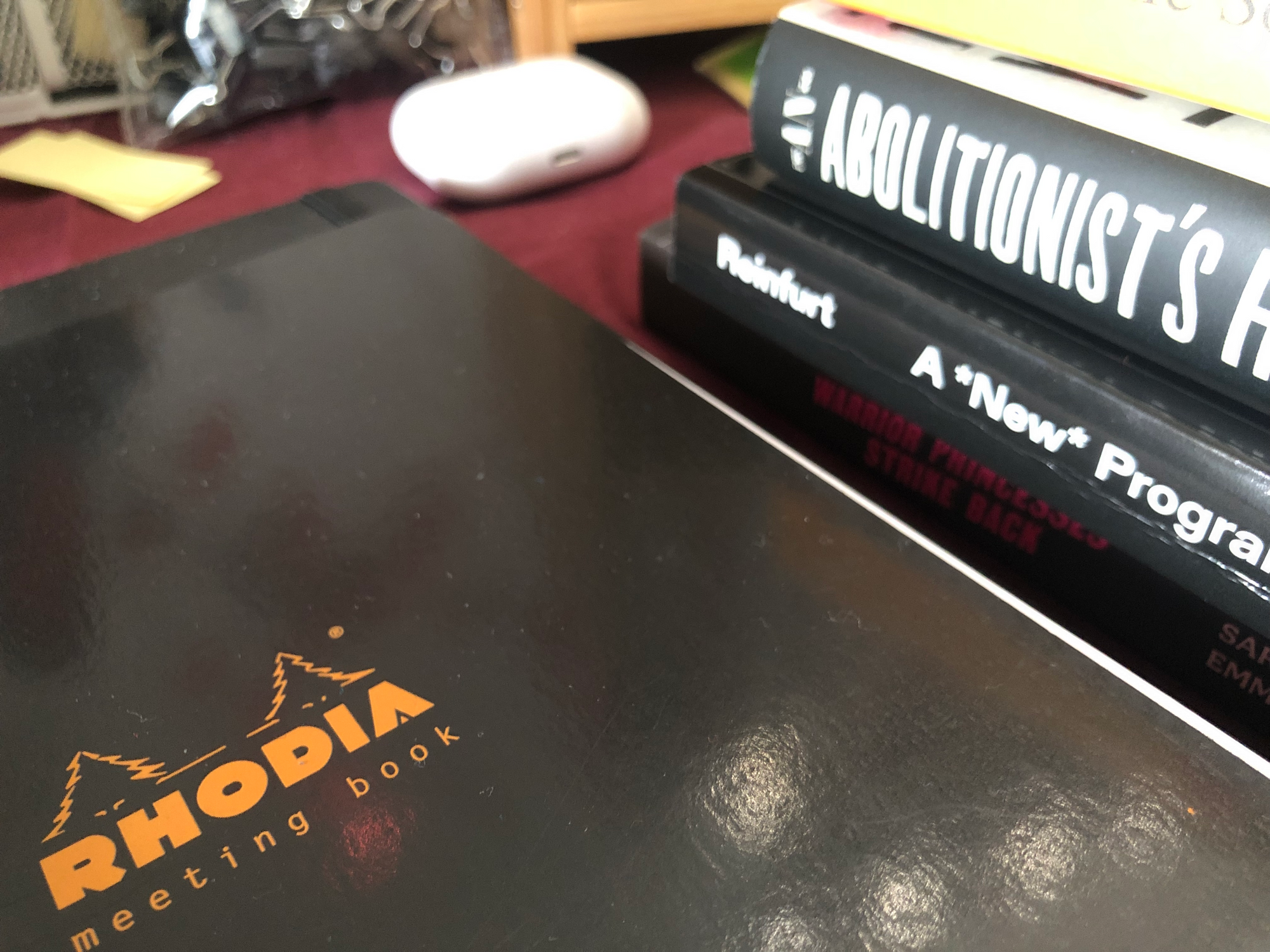 photo of a black Rhodia notebook next to a stack of books, spines partially visible, the top one reading "AN ABOLITIONIST'S", the second "Reinfurt A *New* Program", the third "WARRIOR PRINCESSES STRIKE BACK". AirPods Pro in the background beside a bag of binder clips. taken at the robledo art, strike! headquarters, february 2023.