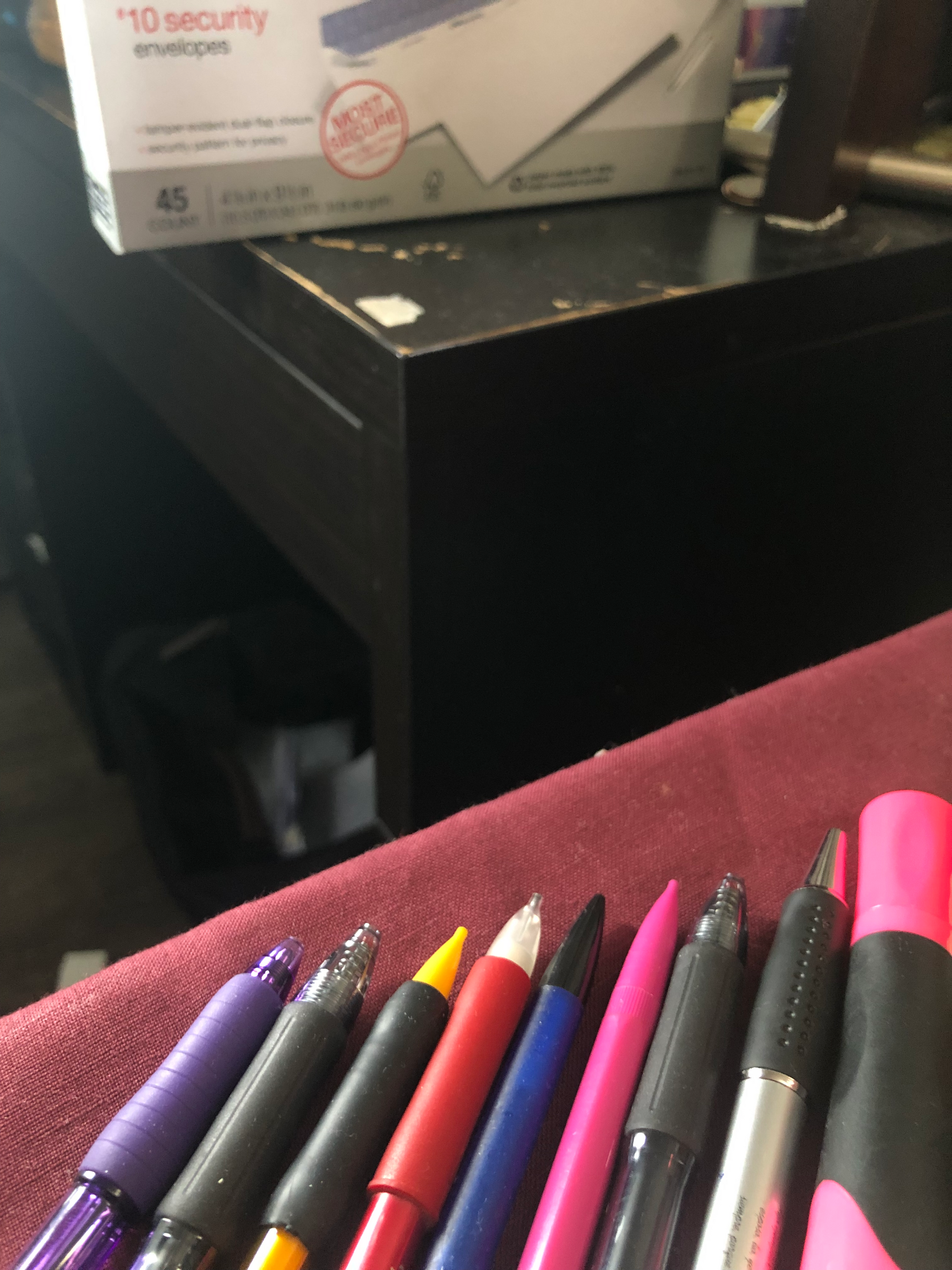photo of a bunch of pens, mechanical pencils, and a pink highlighter on a red tableclothed desk, across from a black desk atop which sits a box of #10 security envelopes. taken at the robledo art, strike! headquarters, february 2023.
