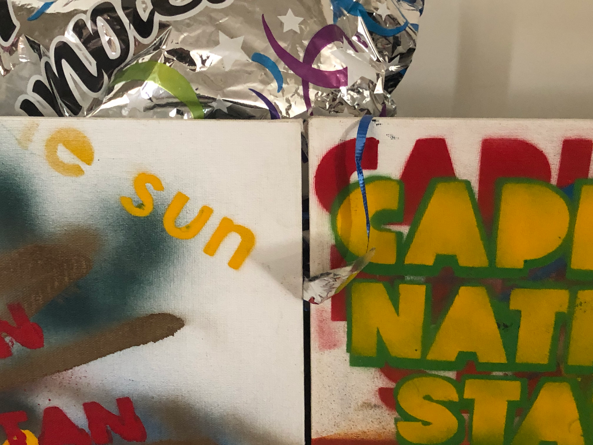 photo of two large canvasses, the top corners visible, the left canvas with dark green and brown streaks, and the letters "e sun" in yellow and "N" and "TAN" in red, the right canvas with the letters "CAPI", "NATI", and "STA" in yellow letters with green outlines, the same letters in red behind, offset. hanging over the canvasses is a blue string attached to a silver celebratory balloon. taken at the robledo art, strike! headquarters, february 2023.
