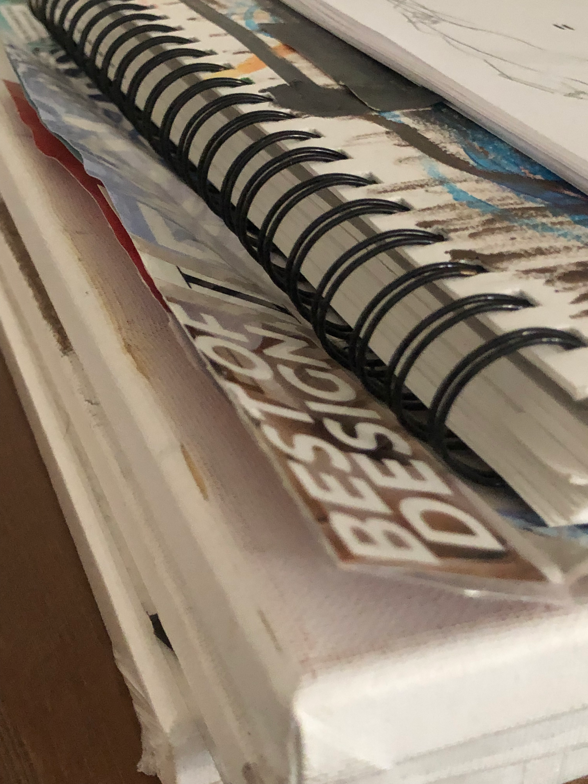 close-up photo of a stack of canvasses, one with a cut-out with the words "BEST OF DESIGN" taped to it, lifting off the canvas, a ringed notebook with an obscured painting and stack of paper on top. taken at the robledo art, strike! headquarters, february 2023.