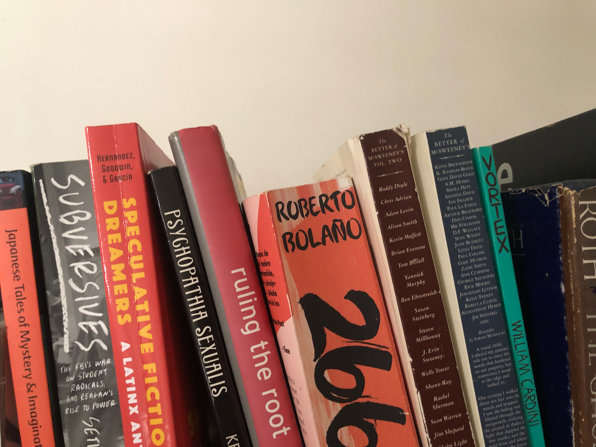 a close-up photo of the tops of spines of a row of books, against a white wall. the readable words on the spines include "Japanese Tales of Mystery & Imagination", "Subversives: The FBI's War on Student Radicals and Reagan's Rise to Power", "Hernandez, Goodwin, & Garcia Speculative Fiction Dreamers A Latinx", "Psychopathia Sexualis", "Ruling the Root", "Roberto Bolaño 266" "The Better of McSweeney's Vol. Two", "The Better of McSweeney's", "Vortex William Cardini" and "Roth". taken at the robledo art, strike! headquarters, february 2023.