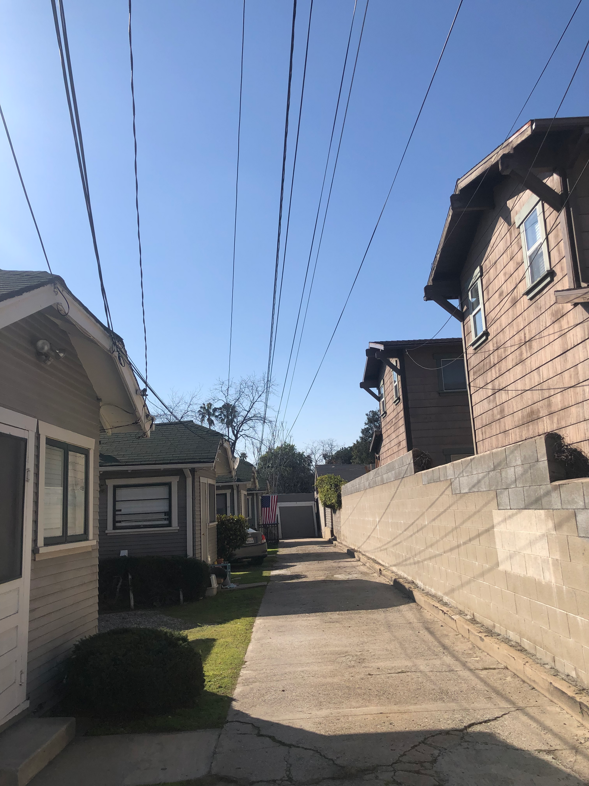 photo of a long paved car-sized path for a row of modest gray houses with dark green roofs, a usa flag hanging at the far house, its garage marking the end of the path. path cracked in the foreground. above stream power lines, casting stark shadows across a worn brick wall separating the homes from two larger light brown houses. taken near the robledo art, strike! headquarters, february 2023.